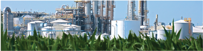 The image depicts a large, industrial ethanol processing facility with grain silos and conveyors with foreground of harvested corn field and a clear, cloudy sky.